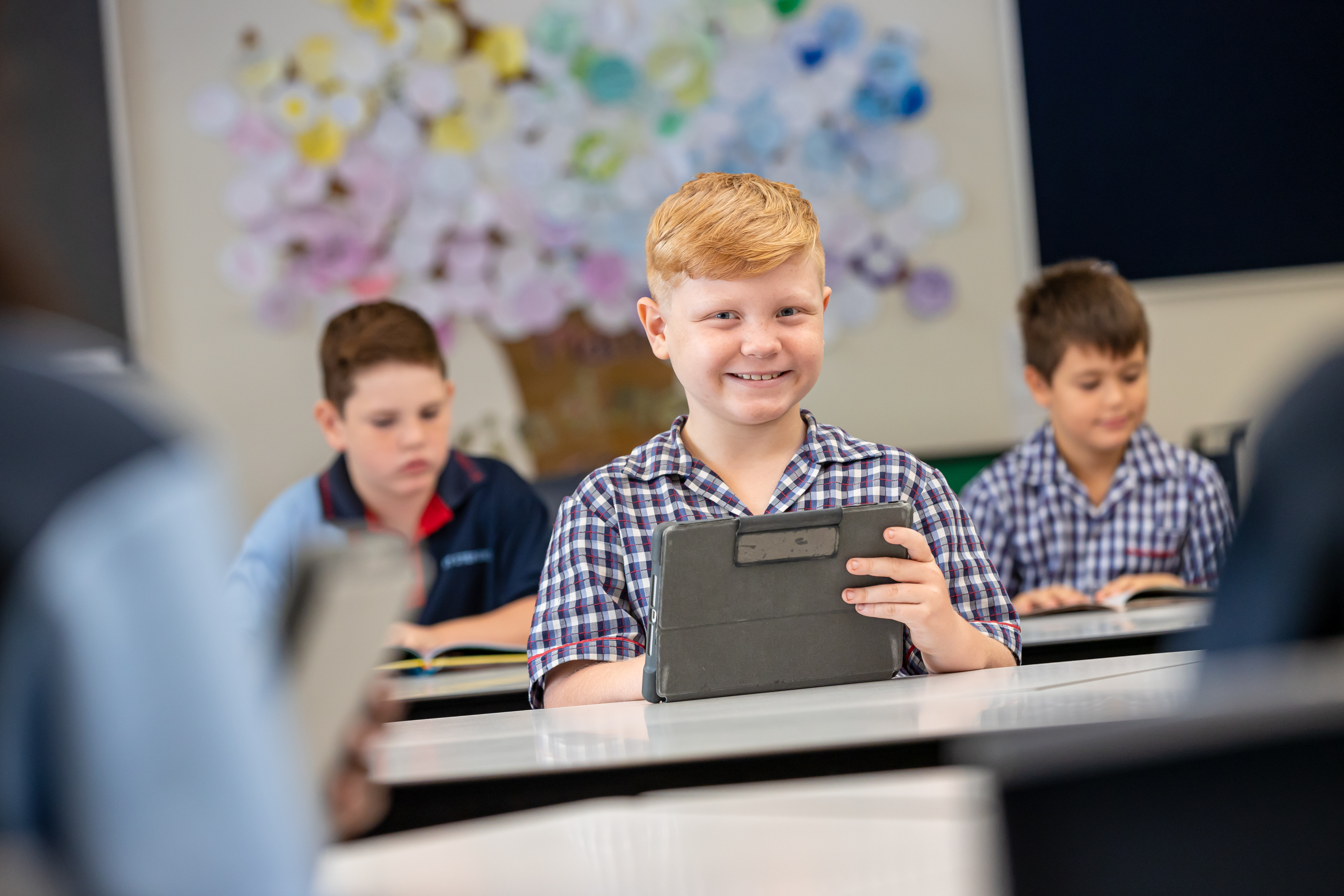A primary aged student is sitting at a desk with an ipad. There are two students seated behind him, reading.